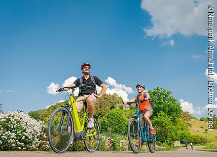 Mit dem E-Bike bequem unterwegs (Naturpark Altmühltal) Zwei Personen fahren bei sonnigem Wetter mit Fahrrädern auf einem Weg in grüner Landschaft.