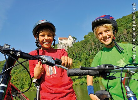 Entspannt am Radweg (Naturpark Altmühltal) Zwei Jungen mit Fahrradhelmen und Fahrrädern vor bewaldetem Hügel und Schloss bei klarem Himmel.