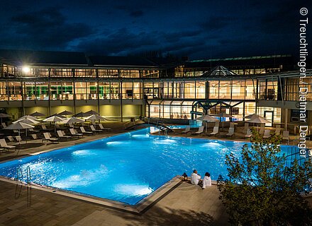 Altmühltherme Treuchtlingen (Naturpark Altmühltal) Beleuchteter Außenpool mit Liegestühlen und Sonnenschirmen vor einem modernen Gebäude bei Nacht.