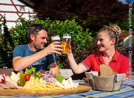 Genuss am Weg (Kipfenberg, Naturpark Altmühltal) Zwei Personen stoßen mit Biergläsern an, davor eine Platte mit Käse, Wurst und Gemüse auf einem Tisch.