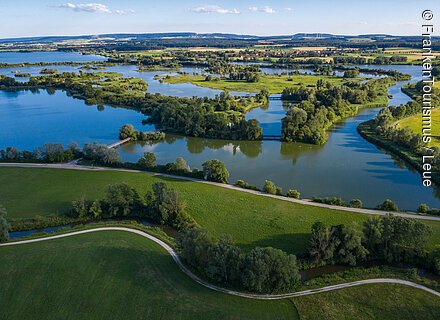 Ferien- und Wasserlandschaften Landschaft mit Seen, Inseln, Bäumen und Wegen unter blauem Himmel, im Hintergrund Felder und Hügel.