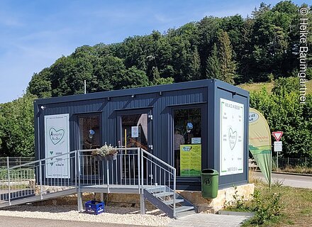 Kiosk am Zeltplatz Hammermühle (Mörnsheim, Naturpark Altmühltal) Kleines Verkaufsgebäude mit Rampe, umgeben von Grün und Wald im Hintergrund.
